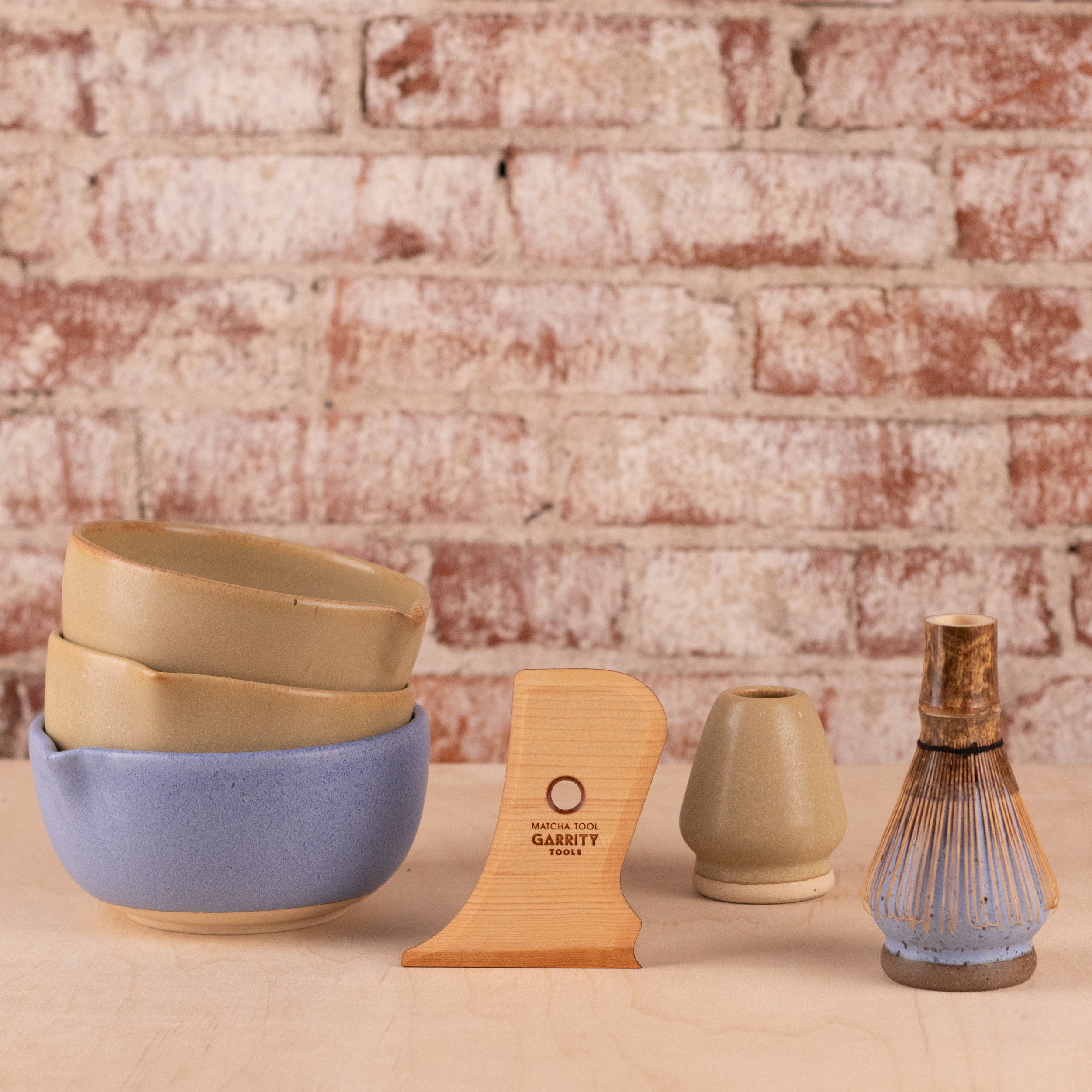 A set of handmade ceramic matcha bowls displayed on a light wood surface against a rustic brick wall background. The set includes a stack of three bowls in beige and blue, a wooden pottery rib engraved with 'GARRITY TOOLS Matcha Tool', a small beige matcha whisk holder, and a blue matcha whisk holder with a bamboo whisk on it.