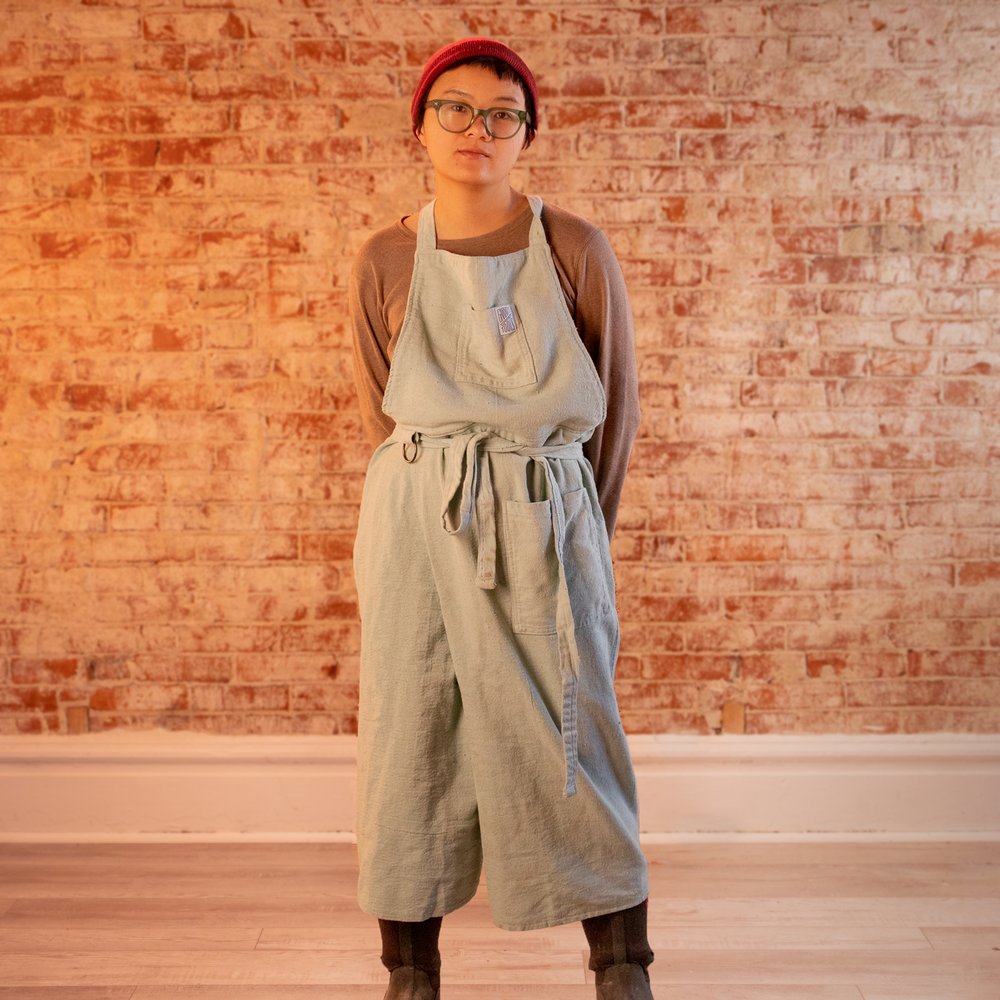 person standing wearing a cotton potter's apron, glasses, and a red hat