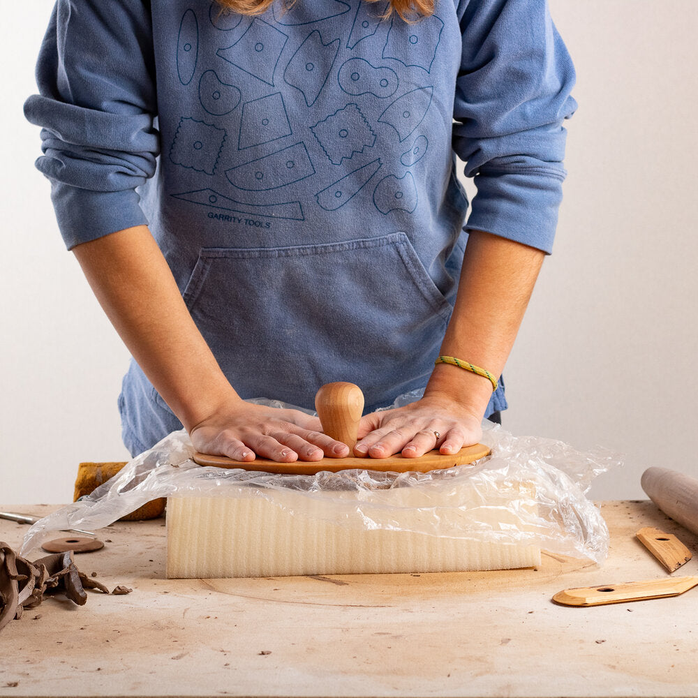 artist using both hands to push wooden anvil top into a clay slab on a piece of foam